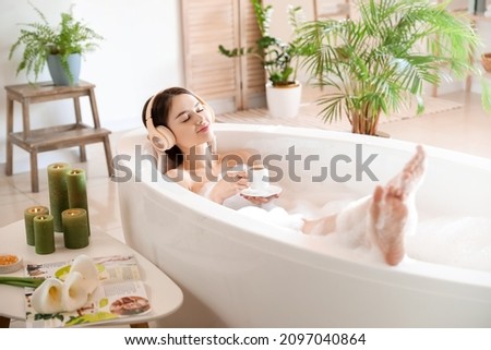 Young woman with headphones and cup of coffee taking bath at home