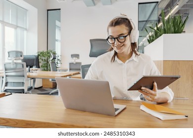 A young woman with headphones, glasses, and a tablet smiles while working on her laptop in a bright, modern office.

 - Powered by Shutterstock