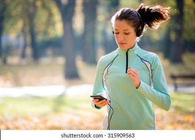 Young woman with headphones jogging in autumn nature and looking to mobile phone - Powered by Shutterstock