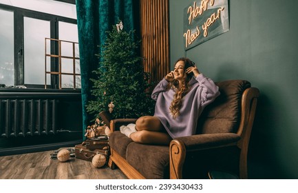 Young woman with headphones listens to music from a vinyl record, sitting on a sofa near a glowing Christmas tree on Christmas Day - Shutterstock ID 2393430433