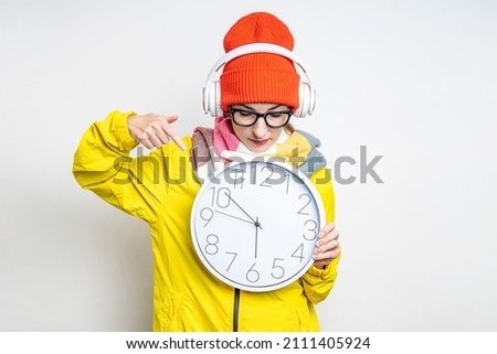 Young woman in headphones pointing her finger at the clock on a light background