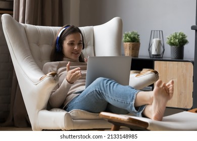 Young Woman In Headphones Relax On Cozy Armchair Put Feet On Footstool, Look At Laptop Screen, Talk To Friend, Share News To Family Living Abroad Through Video Conference App. Virtual Meeting Concept