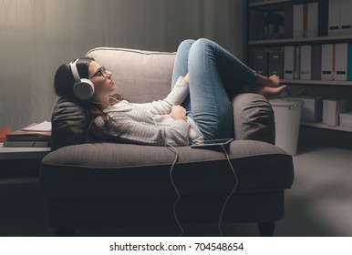 Young Woman With Headphones Relaxing At Home Late At Night, She Is Lying On The Armchair And Listening To Music Using A Tablet