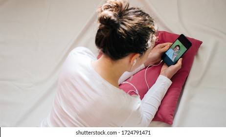 Young woman in headphones using smartphone for video call, gesturing hi to friend lying on bed at home. Girl making facetime video calling with smartphone.