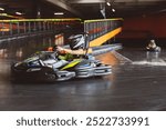 Young woman in helmet driving a go-kart on an indoor track, racing with another kart in the background.