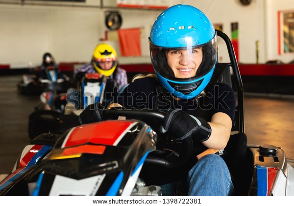 Young woman in helmet and other people\
driving cars for racing in sport club\
indoor