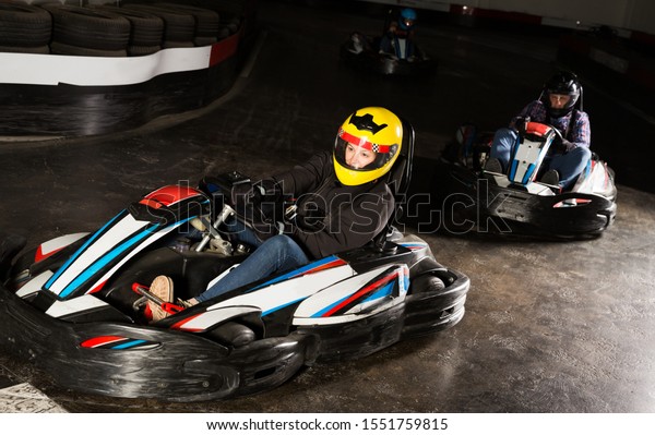 Young woman in helmet and other people\
driving cars for karting in sport club\
indoor