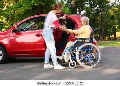 Young Woman Helping Disabled Grandmother In Wheelchair To Get Into Car Outdoors