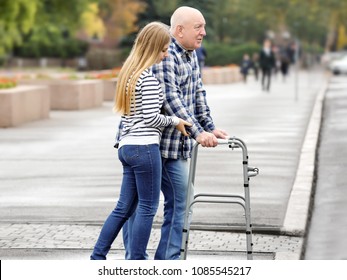 Young Woman Helping Elderly Man With Walking Frame To Cross The Road