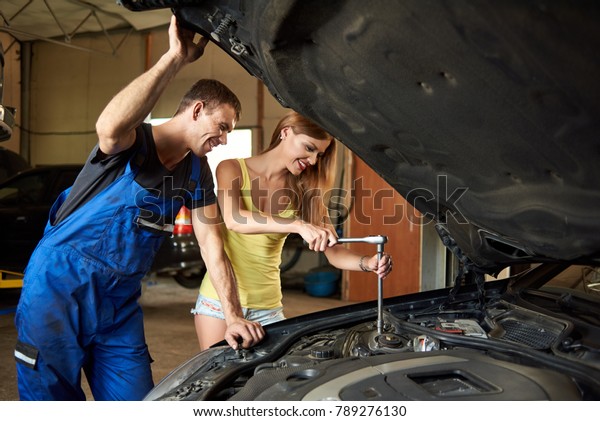 Young woman helps an auto mechanic repair a car\
in a garage. The girl unscrews the front right-hand stand. The guy\
holds the hood cover by\
hand