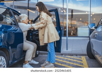 A young woman helps an elderly woman out of a car in a parking lot, demonstrating care and support. The setting suggests a shopping or healthcare environment. - Powered by Shutterstock