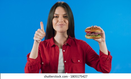 Young Woman In Her 20s On A Blue Background In Studio, Showing That The Hamburger In Her Hand Is Delicious.