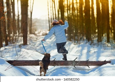 Young Woman With Her Dog Jumping Over A Log In Snowy Winter