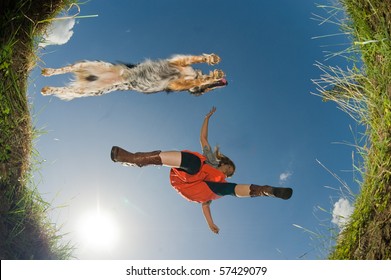 Young Woman And Her Dog Jumping Over A Creek, Shot From Below.