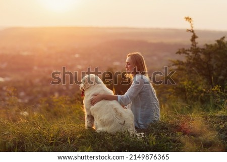 A young woman and her dog sitting on the hill in the countryside on a summer evening. A lady and her faithful friend enjoying beautiful landscape at summer sunset.