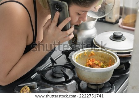 young woman in her kitchen talking on her cell phone, while uncovering a pot where she is preparing healthy food.