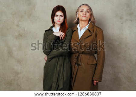 A young woman with her mother are standing in identical cloaks against a gray fabric background                  