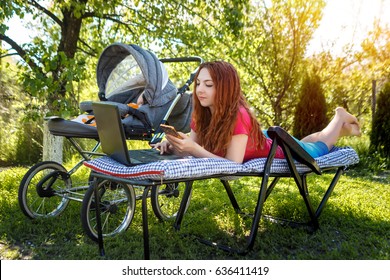 Young Woman With Her Newborn Baby Lying On The Soft Armchair With Laptop And Looking At The Phone. Summer Day. Mother With Child Outdoors. Motherhood.