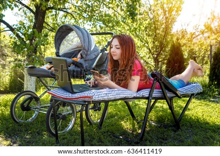 Young woman with her newborn baby lying on the soft armchair with laptop and looking at the phone. Summer day. Mother with child outdoors. Motherhood.