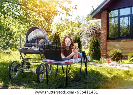 Young woman with her newborn baby lying on the soft armchair with laptop and doing selfei. Summer day. Mother with child outdoors. Motherhood.