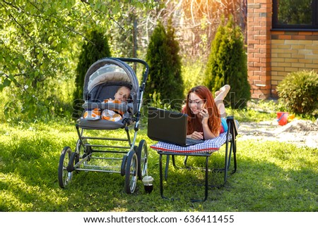 Young woman with her newborn baby lying on the soft armchair with laptop and talking on phone. Summer day. Mother with child outdoors. Motherhood.