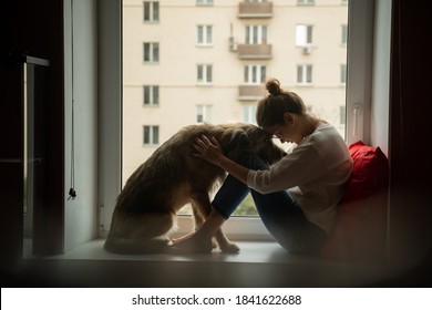 A young woman with her shaggy dog at home, sits on a windowsill overlooking the city. Self-isolation, quarantine. Love for pets, joy and tenderness