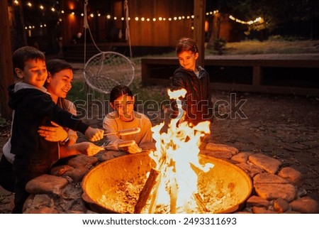 A young woman with her three children is frying marshmallows on the fire. An attractive mom with her son and daughters are having fun in the backyard. A happy family outdoors at home by the campfire
