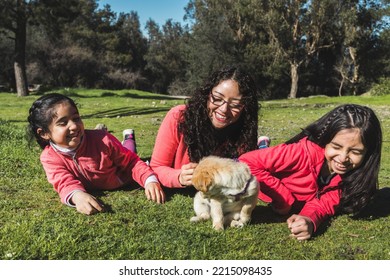 Young Woman With Her Two Daughters, Lying On The Grass And Playing With A Golden Retriever Puppy In The Park