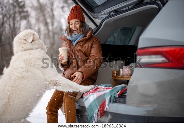 Young woman with her white dog drink a coffee in\
car trunk near a snowy forest, traveling by car during winter\
holidays. High quality\
photo