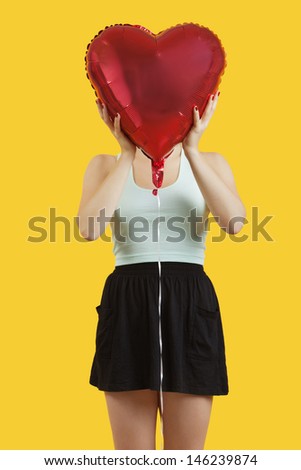 Young woman hiding behind heart shaped balloon over yellow background