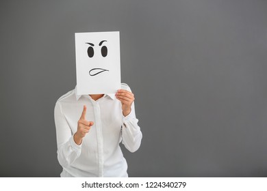 Young Woman Hiding Face Behind Sheet Of Paper With Drawn Emoticon On Grey Background