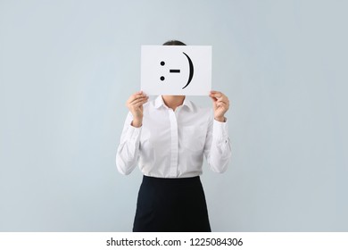 Young Woman Hiding Face Behind Sheet Of Paper With Drawn Emoticon On Light Background