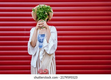 Young Woman Hiding Her Face With Bouquet Of Basilic, Having Net Shopping Bag With Tomatoes Standing Over Red Background Outdoor. Concept Of Sustainability, Healthy Food And Eco-friendly Lifestyle.