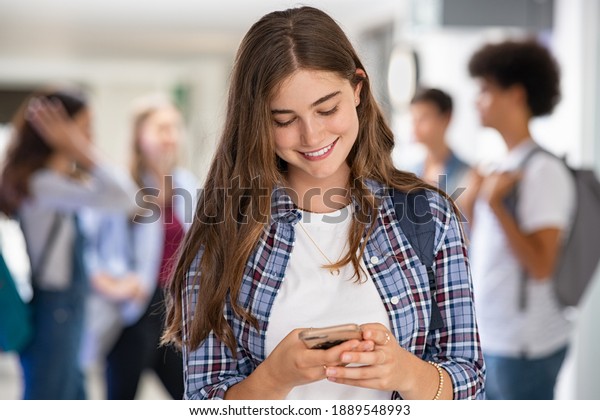 Young woman in\
high school campus using smartphone. Smiling university student\
texting on mobile phone standing in hallway. Happy college girl\
messaging on smart phone, using\
app.
