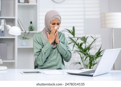 Young woman in hijab working from home and using tissue while sneezing at desk with laptop and tablet in modern, bright office setting - Powered by Shutterstock