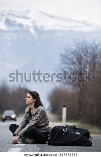 Young woman hitchhiking on countryside\
road.Traveler woman sitting alone along the road.Pretty young woman\
tourist hitchhiking.Left alone on the road and\
lost