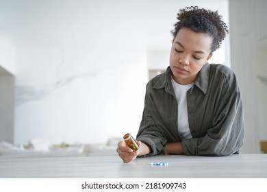 Young woman hold bottle with pills read medicine instruction before take meds. Pensive girl holding medicines thinking of taking medications, doubt whether to take tablets, sitting at table. - Shutterstock ID 2181909743