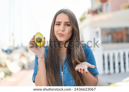 Young woman holding an avocado at outdoors making doubts gesture while lifting the shoulders
