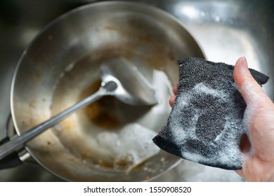 Young Woman Holding Black Metal Sponge With White Bubble Foam From Dishwashing Liquid. Prepare To Scrub Dirty Stainless Pan In Sink. Cleaning Kitchenware. Hard Stain. Copy Space.