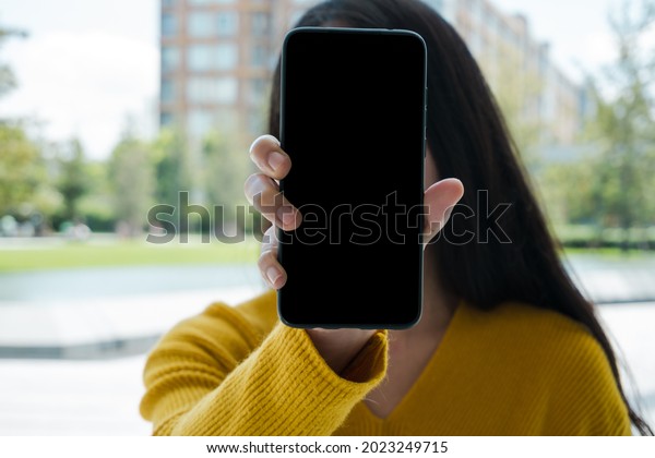 Young woman holding a\
black phone showing off. In the background are buildings and trees,\
soft blur.