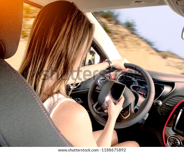 Young woman holding blank screen cell phone\
while driving car. Female driver hand on steering wheel &\
checking out her smartphone in moving vehicle. Don\'t text and drive\
concept. Close up,\
background.
