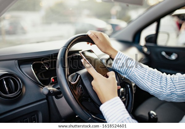 Young woman holding blank screen cell\
phone, driving car. Female business woman driver hand on steering\
wheel & checking smartphone in moving vehicle. Don\'t text and\
drive concept. Close\
up,background