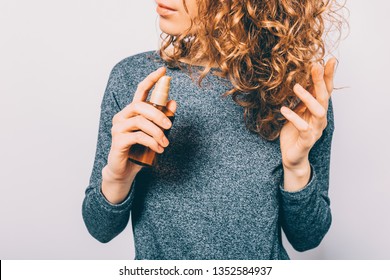 Young woman holding bottle with nutritional oil applying on her curly brown hair. Female's hands using cosmetic serum to prevent split ends.