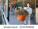 Young woman holding bucket of hay feed for horse in stable