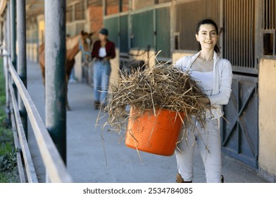 Young woman holding bucket of hay feed for horse in stable - Powered by Shutterstock