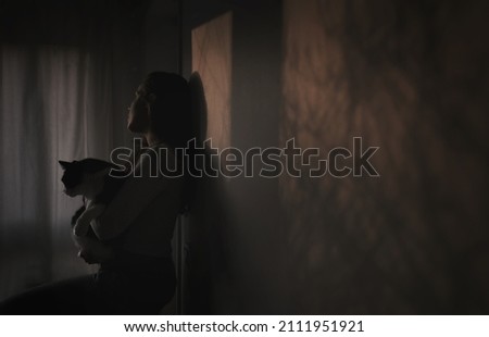 Young woman holding a cat in the arms in a dark room looks away towards a window.
