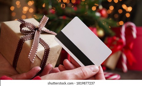 Young Woman Holding A Credit Card And A Gift Box Against The Background Of Christmas Decor And Gifts, Close-up. Christmas And New Year Shopping On The Internet, Payment By Credit Card.