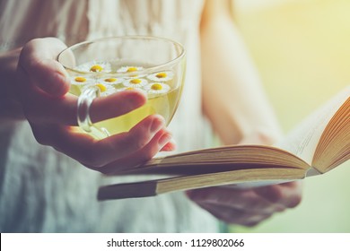 Young Woman Holding Cup Of Chamomile Tea And Reading Open Book