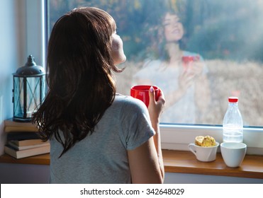 Young  Woman Holding Cup Of Coffee Sitting Home By The Window. Face Reflection On The Glass