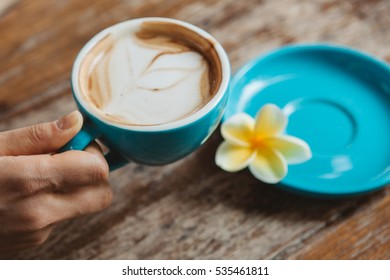 Young woman holding a cup of morning coffee cappuccino served on wooden table with plate and frangipani flower